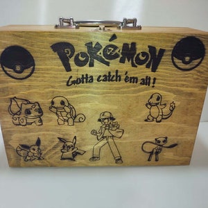 Könnte beinhalten: Eine Holzkiste mit einem Verschluss und einem Pokemon-Motiv. Die Kiste ist mit einem Pokemon-Logo, dem Spruch "Gotta catch 'em all!" und mehreren Pokemon-Charakteren verziert, darunter Pikachu, Bisasam, Glumanda, Schiggy und ein Trainer.