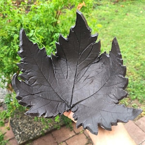 May include: A dark gray, leaf-shaped decorative item with detailed veining and a textured surface. The leaf has a jagged edge and is held against a background of green foliage and brick pavers.