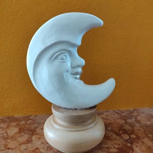 Pode incluir: Uma escultura de lua crescente de cerâmica branca com um rosto sorridente, montada em uma base de madeira.