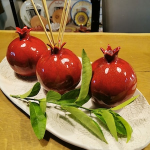 Pode incluir: Três difusores de cerâmica vermelha em forma de romã com varetas de madeira em um prato oval branco com folhas verdes.