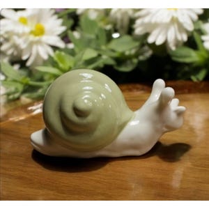 May include: A white ceramic snail figurine with a green shell. The snail is in a crawling position with its shell facing forward.