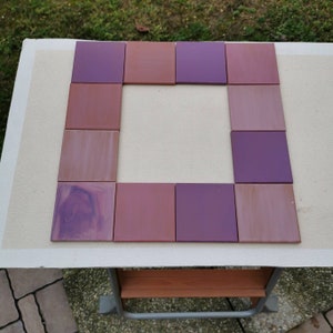 Könnte beinhalten: Ein Satz von 12 quadratischen Fliesen in Violett- und Rosatönen, die in Form eines Rahmens angeordnet sind. Die Fliesen bestehen aus einem glatten, glänzenden Material.