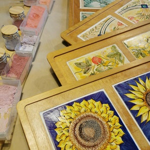 Może przedstawiać: Ręcznie robione płytki ceramiczne z kwiatowymi wzorami, w tym słoneczniki i truskawki, w drewnianych ramach. W pobliżu ustawiono r&oacute;żne słoiki i pojemniki z kolorowymi proszkami i pigmentami, co sugeruje pracownię artystyczną lub warsztat.