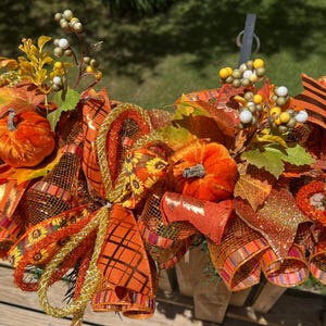 May include: A fall-themed decorative arrangement featuring orange pumpkins, autumn leaves, and decorative ribbons. The arrangement includes a variety of textures and colors, with accents of gold and green. Perfect for seasonal home decor.