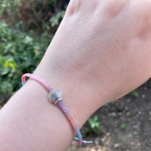 May include: A pink, blue and white friendship bracelet with a silver seashell charm.