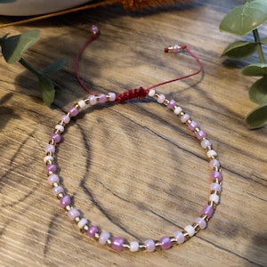 May include: A beaded bracelet with a sliding knot fastening. The bracelet features small, round beads in shades of pink, white, and gold. The cord is a deep red colour.