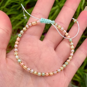 May include: A pastel-coloured beaded bracelet with pink, yellow, and green beads, and gold-coloured accents. The bracelet has a light blue macrame knot and string closure. The beads are small and evenly spaced. A delicate piece of jewellery.
