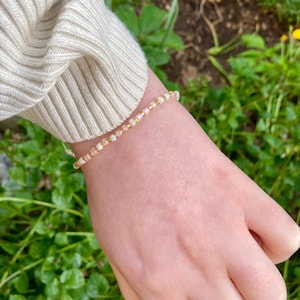 May include: A delicate beaded bracelet with small white, pink, and gold beads. The bracelet is worn on a wrist, with a cream-coloured sweater sleeve in the background. The bracelet is a dainty piece of jewellery, perfect for everyday wear.