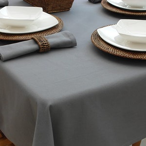 May include: A grey tablecloth with a woven placemat and white bowl on a white plate. The placemat is made of natural materials and is brown in colour.
