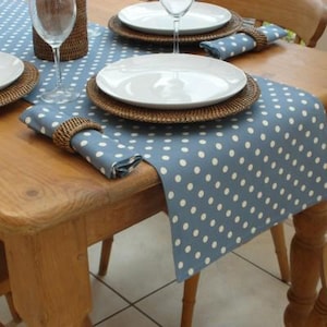 May include: A dining table set with a blue polka dot table runner, white plates, woven placemats, and matching napkins. Wine glasses are placed on the table. The table is made of light-coloured wood.