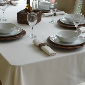 May include: A white tablecloth with a wicker charger plate, a white bowl, and a white napkin ring. The table is set for a formal dinner with glasses and cutlery.