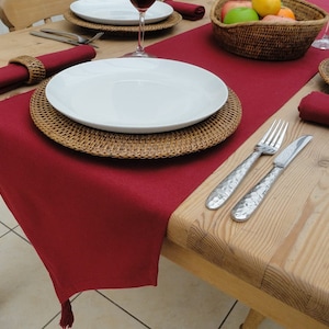 May include: A red table runner with a tassel fringe, set on a wooden table with a woven charger, white dinner plate, and cutlery. A bowl of fruit and a glass of wine are also visible.