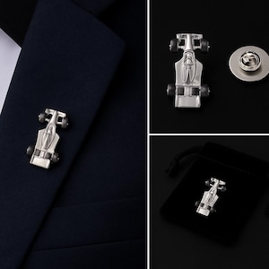 Può includere: Una spilla da bavero in metallo color argento a forma di auto da corsa di Formula 1. La spilla presenta un design aerodinamico con quattro piccole ruote nere. È mostrata fissata su un tessuto blu navy scuro e include una chiusura rotonda.