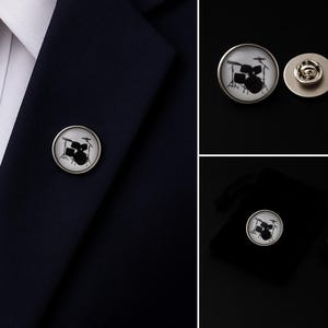 Op de afbeelding: Een ronde reversspeld met een zwarte silhouet van een drumstel tegen een witte achtergrond. De speld heeft een zilverkleurig metalen frame en een standaard sluiting, afgebeeld bevestigd aan een donkerblauw kledingstuk van stof.