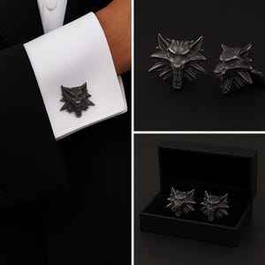 Può includere: Un paio di gemelli in metallo scuro con un design a testa di lupo scolpita, orecchie appuntite e bocca aperta. I gemelli sono mostrati fissati a un polsino di camicia bianco e anche all'interno di una scatola nera rettangolare.