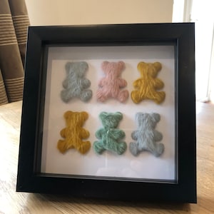 May include: A framed artwork featuring six fuzzy teddy bears in different colors, including blue, pink, yellow, green, and gray. The bears are arranged in a grid pattern on a white background.