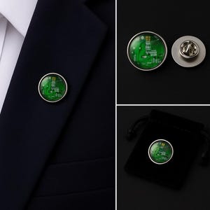 May include: A circular lapel pin featuring a green printed circuit board pattern encased in a silver-toned metal frame. The pin includes a matching silver-toned butterfly clutch backing and is displayed alongside a small black velvet drawstring pouch.