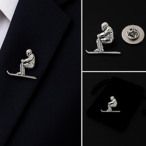 May include: A silver-toned metal lapel pin shaped like a skier in a crouched position, wearing a helmet and goggles, holding a ski pole while standing on a single ski. The pin features a detailed, textured finish and includes a circular metal backing.