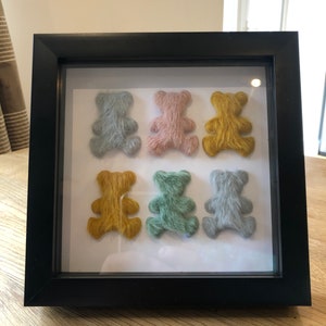 May include: A framed display of six small, fuzzy teddy bears in a variety of colors, including blue, pink, yellow, and green. The bears are arranged in two rows of three.