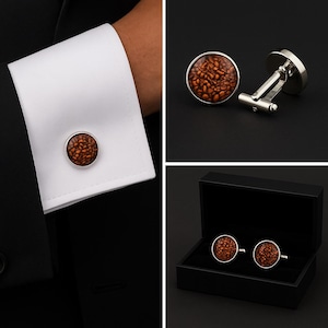 May include: Round silver-tone cufflinks featuring a textured, amber-colored inlay with a pattern resembling small, clustered coffee beans. The set includes one cufflink attached to a white shirt cuff and a black presentation box for storage.