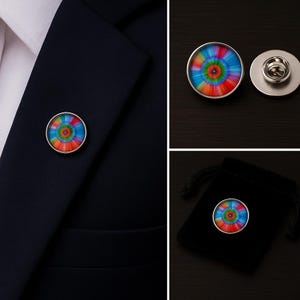 May include: A circular lapel pin featuring a vibrant, multi-colored radial pattern with shades of blue, green, yellow, orange, and red. The pin has a metallic silver-toned frame and includes a matching metal clutch back and a small black pouch.