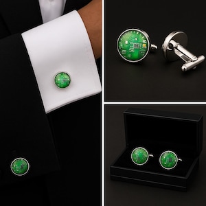 May include: A pair of round silver-toned cufflinks featuring a green circuit board pattern under a glass dome. The cufflinks are shown attached to a white shirt cuff and also displayed inside a black rectangular presentation box.