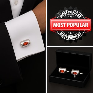 Può includere: Un paio di gemelli color argento dal design cilindrico con un inserto rettangolare arancione al centro. I gemelli sono mostrati fissati a un polsino di camicia bianco e anche all'interno di una scatola rettangolare nera.