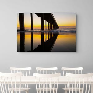 Puede incluir: Un cuadro rectangular que muestra la silueta de un largo muelle sobre el océano al atardecer. El cielo presenta un degradado de naranja y amarillo, con la estructura oscura del muelle reflejada claramente en la arena húmeda bajo el horizonte.