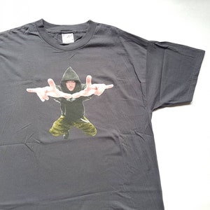 May include: A dark gray t-shirt with a graphic print of a person wearing a black hooded sweatshirt and camouflage pants. The person is making a hand gesture with both hands.