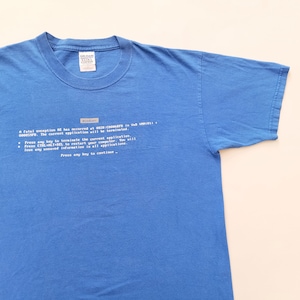 May include: A blue t-shirt featuring a printed Windows error message. The text includes the word "Windows" and a series of lines describing a fatal exception. The shirt is made of a soft material.