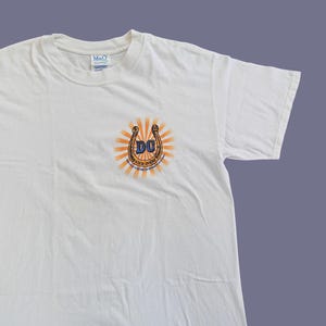 May include: White t-shirt with a graphic of a horseshoe with the letters "DC" inside and the text "Dixie Chicks" below.