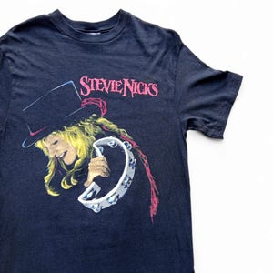 Könnte beinhalten: Marineblaues T-Shirt mit einem Aufdruck von Stevie Nicks mit Zylinder und Tamburin. Der Text "Stevie Nicks" ist rosa gedruckt. Das Shirt hat einen Rundhalsausschnitt und kurze Ärmel.