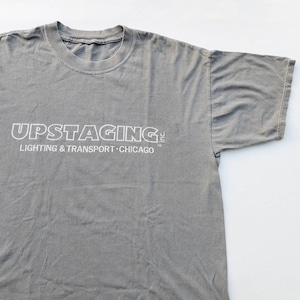 Op de afbeelding: Een grijs T-shirt met de witte tekst "UPSTAGING INC. LIGHTING & TRANSPORT - CHICAGO" gedrukt op de voorkant.