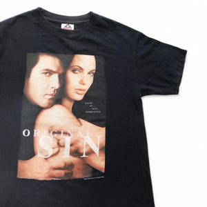 以下が含まれることがあります： 「オリジナル・シン」の映画のアンジェリーナ・ジョリーとアントニオ・バンデラスの写真がプリントされた黒いTシャツ。Tシャツには「Lead us into temptation」という文字がプリントされています。