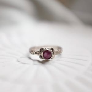 May include: A silver ring with a delicate design and a small, oval-shaped, dark pink gemstone.