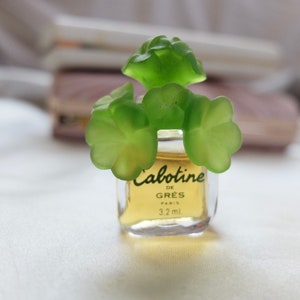 Peut inclure: Un petit flacon de parfum Cabotine de Gres avec un bouchon en forme de trèfle vert. Le flacon est transparent et le liquide à l'intérieur est jaune. L'étiquette sur le flacon indique "Cabotine de Gres Paris 3.2 ml".