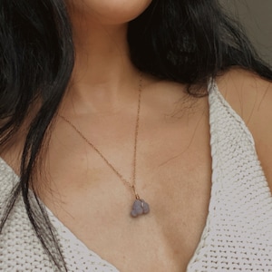 May include: A woman wearing a white knitted top with a gold chain necklace and a purple stone pendant.