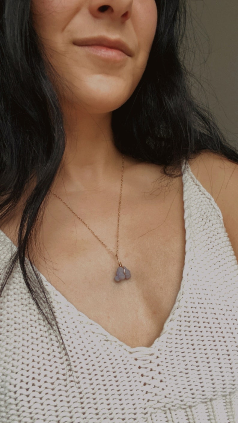May include: A woman wearing a white knitted top with a gold chain necklace and a purple stone pendant.