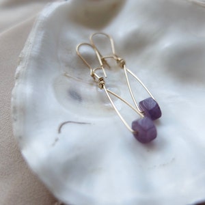 May include: A pair of gold earrings with purple gemstone beads. The earrings are hanging from a white seashell.