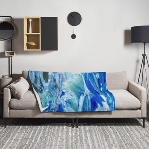 Könnte beinhalten: Eine blaue und weiße abstrakte Wolldecke mit weicher, flauschiger Textur. Die Decke liegt auf einem grauen Sofa in einem Wohnzimmer.