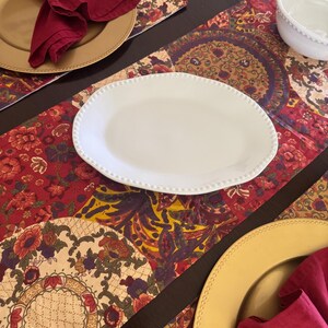 May include: A table setting featuring a white oval plate with a beaded rim, two gold charger plates with deep red cloth napkins in silver rings, and a white bowl, all arranged on a runner with a multi-colored floral and geometric pattern.