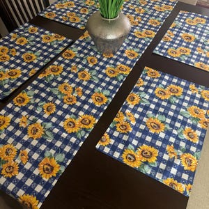 May include: A set of rectangular placemats and a matching table runner featuring a navy blue and white gingham check pattern decorated with vibrant yellow sunflowers and green leaves, arranged on a dark brown wooden dining table surface.