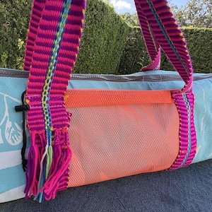 May include: A light blue cylindrical yoga mat bag featuring a white whale graphic, a bright orange mesh exterior pocket, and vibrant magenta woven straps with multi-colored fringe accents. The bag includes a gray zipper closure along the top.