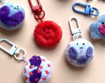Puede incluir: Cinco llaveros de peluche con diferentes diseños. Cada llavero presenta un personaje redondo y peludo con una cara y un color diferente. Los llaveros están unidos a anillos de metal.