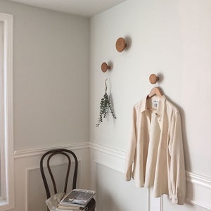 Könnte beinhalten: Drei Wandhaken aus hellem Holz sind an einer weißen Wand montiert. Ein weißes Hemd hängt am unteren Haken. Ein Zweig Grün hängt am mittleren Haken.