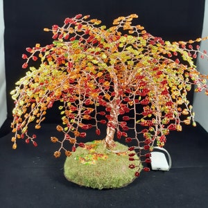 May include: A handcrafted wire tree sculpture with a copper trunk and branches, adorned with colourful beads in shades of red, orange, and yellow, resembling autumn foliage. The tree sits on a moss-covered base, creating a decorative accent.