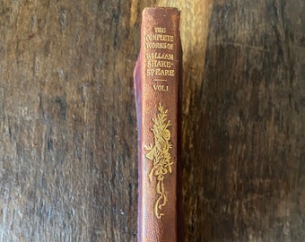 Peut inclure: Un dos de livre en cuir marron intitulé The Complete Works of William Shakespeare Vol. I avec des lettres dorées en relief et un emblème floral décoratif. Le mot Nelson est imprimé en doré au bas du dos du livre.