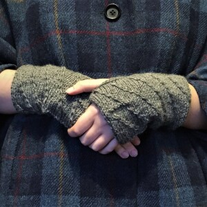 以下が含まれることがあります： グレーのニットの指なし手袋のペア。手袋は、格子縞のシャツを着た人の手に折り畳まれて保持されています。