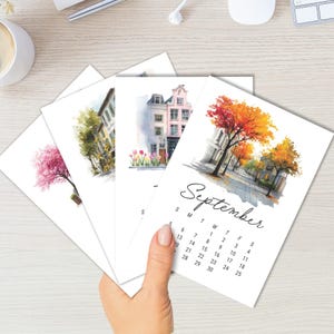 Puede incluir: Un conjunto de cuatro tarjetas de calendario rectangulares con ilustraciones en acuarela de edificios de estilo europeo y &aacute;rboles estacionales. La tarjeta superior muestra el mes de septiembre con una escena callejera con follaje oto&ntilde;al naranja y amarillo.