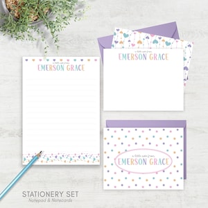 May include: A stationery set featuring a notepad, notecards, and envelopes. The set includes the text "EMERSON GRACE" in pastel colors, with heart and dot patterns. The notepad has lined pages and a blue pencil.